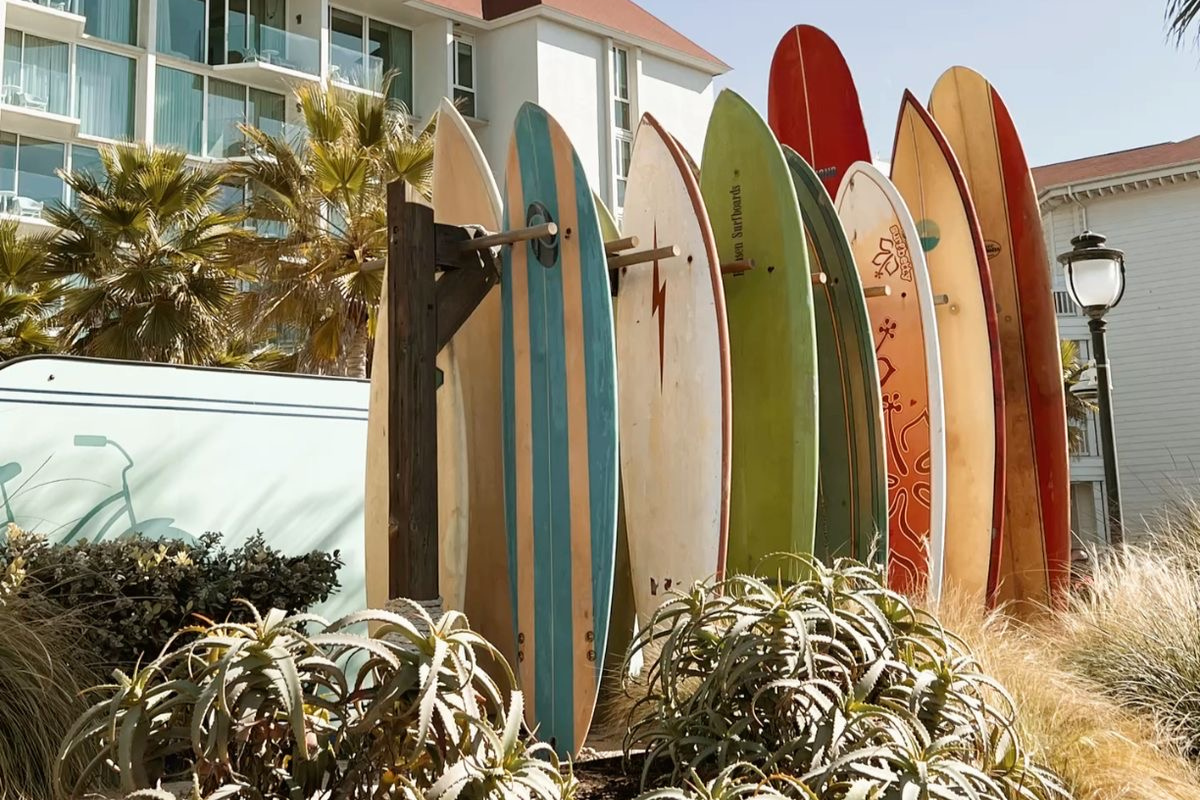 A row of colorful, vintage surfboards standing upright in an outdoor wooden rack near a coastal resort building with palm trees.