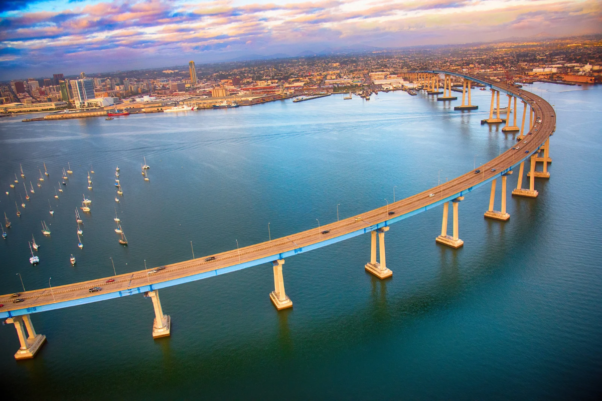 An aerial view of the long, curving San Diego-Coronado Bridge sweeping over calm bay water toward the city skyline during sunset.