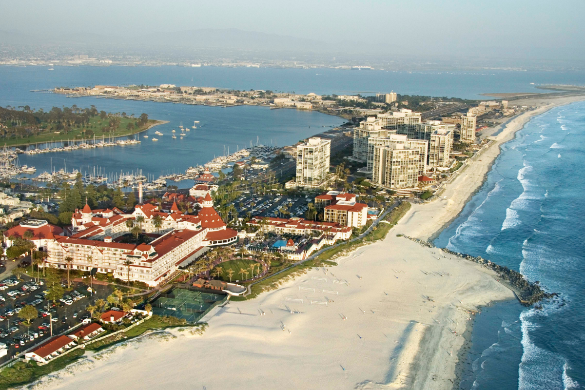 Aerial view of the Hotel del Coronado along a wide sandy beach and ocean, with high-rise buildings and a marina in the background.
