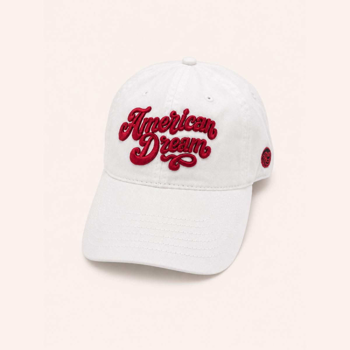 High-angle front view of a clean white cotton baseball cap against a neutral off-white background. The crown features bold, 3D puff red embroidered script reading "American Dream" in a retro cursive font, with a small red circular logo visible on the left side panel.