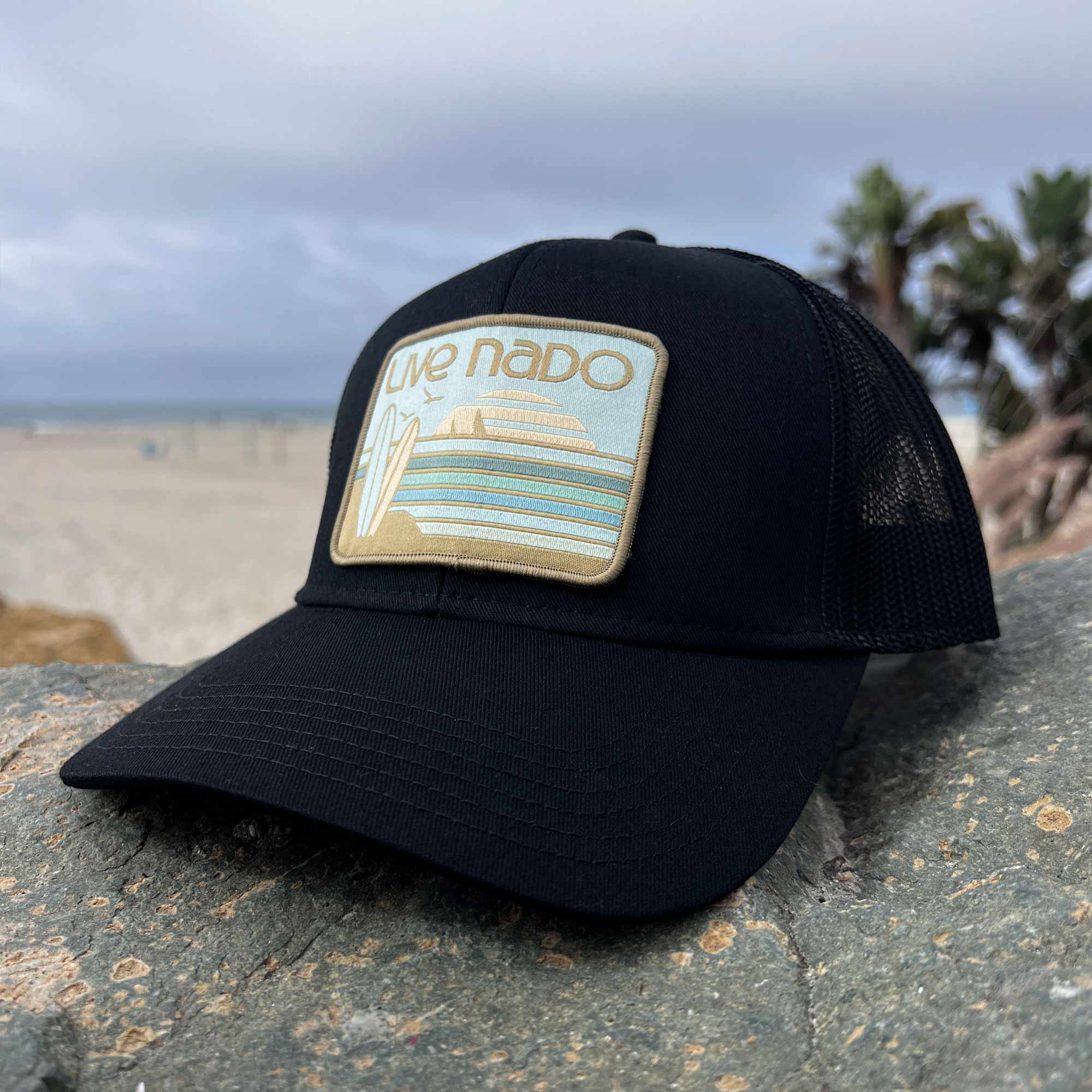 Close up of black Coronado trucker hat with neutral green tones, surfboards and beautiful sunset