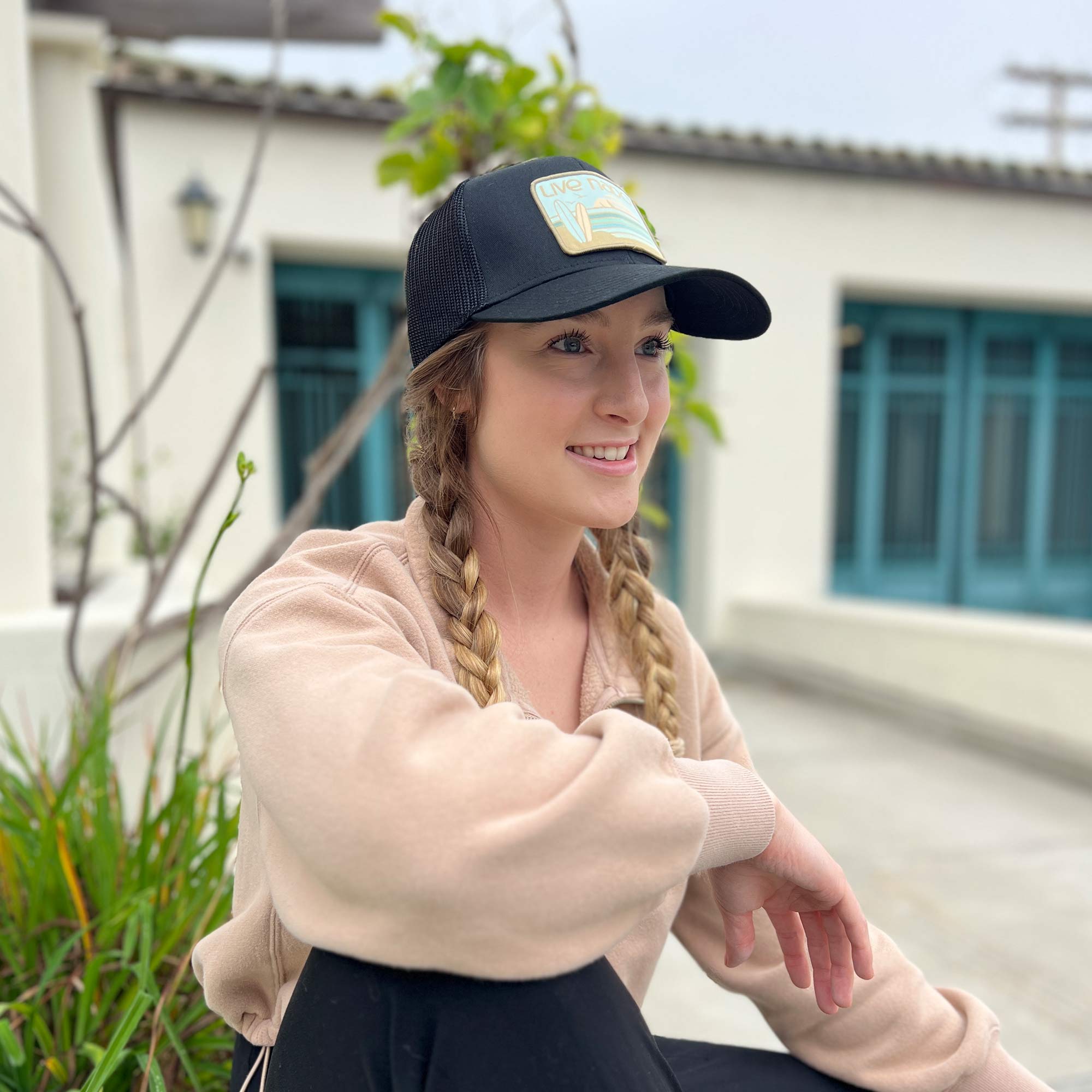 Woman wearing black mesh trucker hat from Coronado. Has an embroidered patch with neutral greens, surfboards and a sunset.
