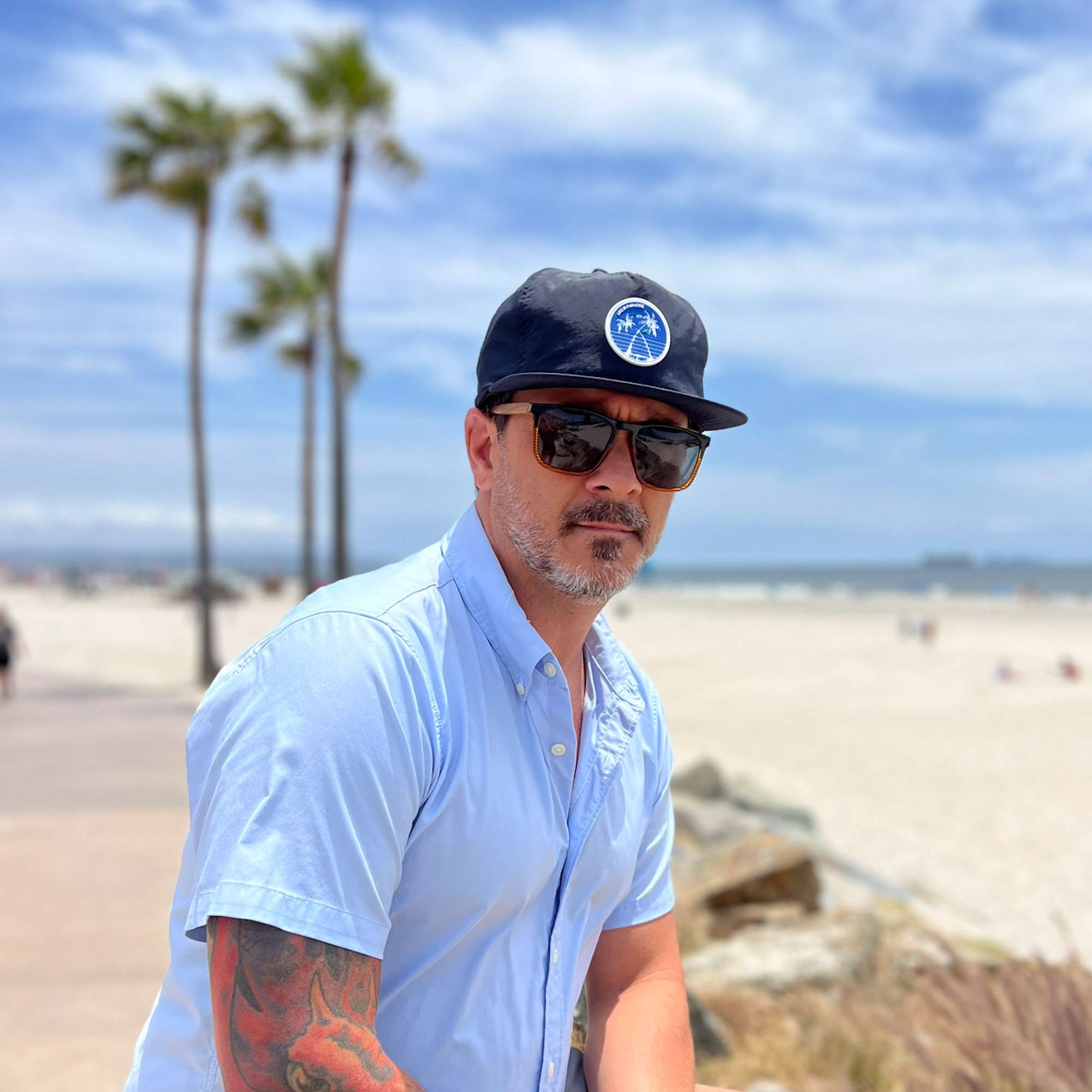man wearing navy, flat brim unstructured hat with blue and white palm tree rubber patch standing near Coronado beach