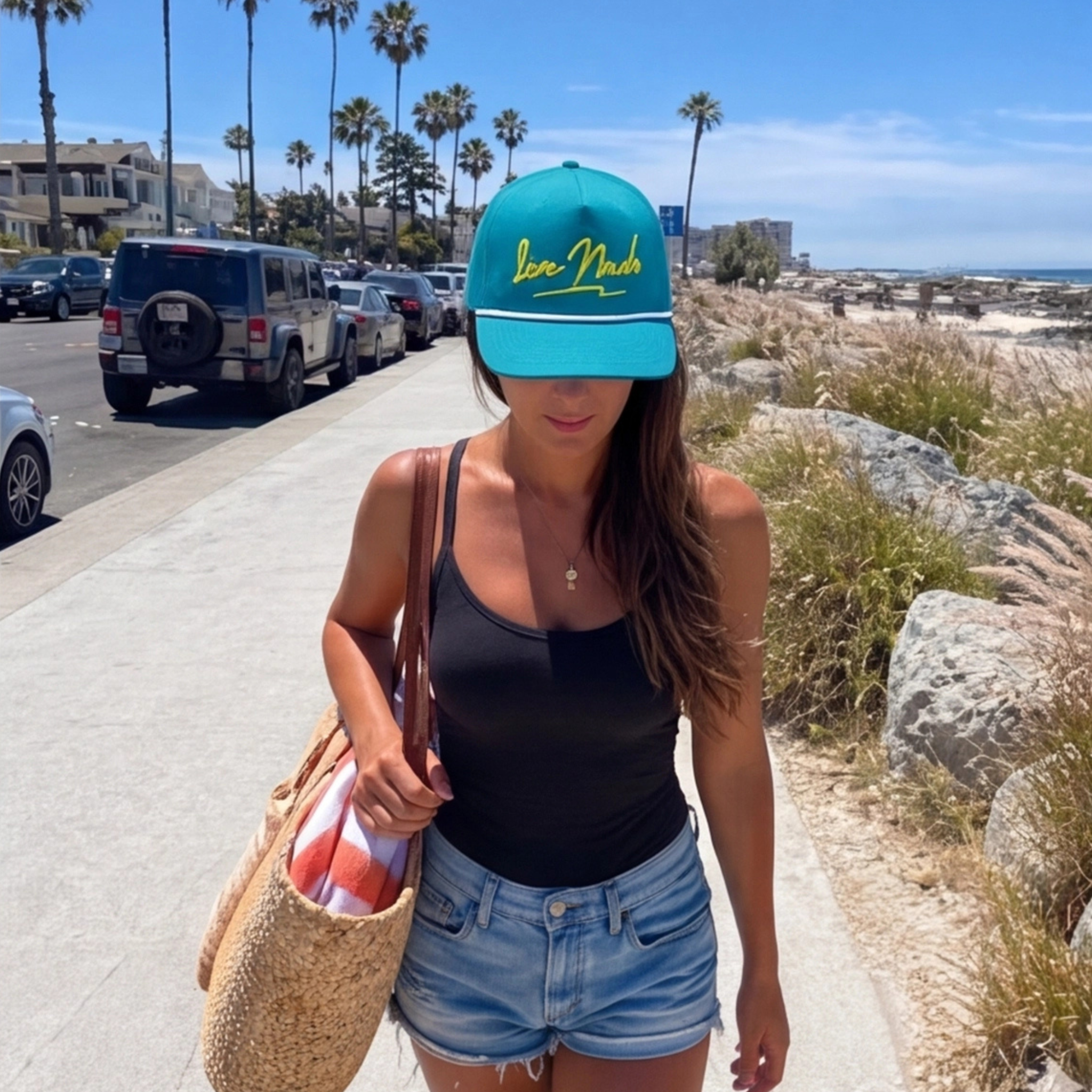 Woman wearing a blue cap with yellow text, black tank top, and denim shorts walking on a sidewalk near palm trees and the ocean.