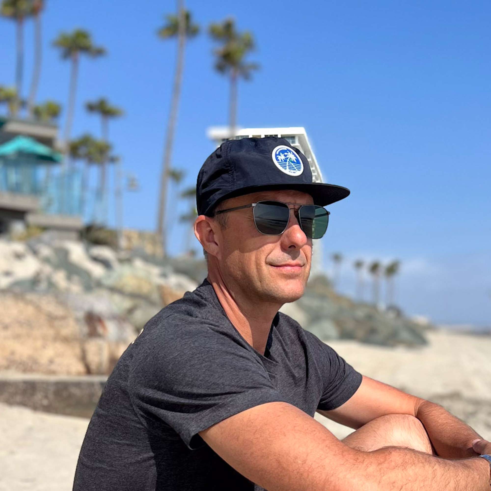 man sitting near hotel del wearing navy blue flat brim surf cap with silicone palm tree patch. Patch represents beautiful Coronado beaches.
