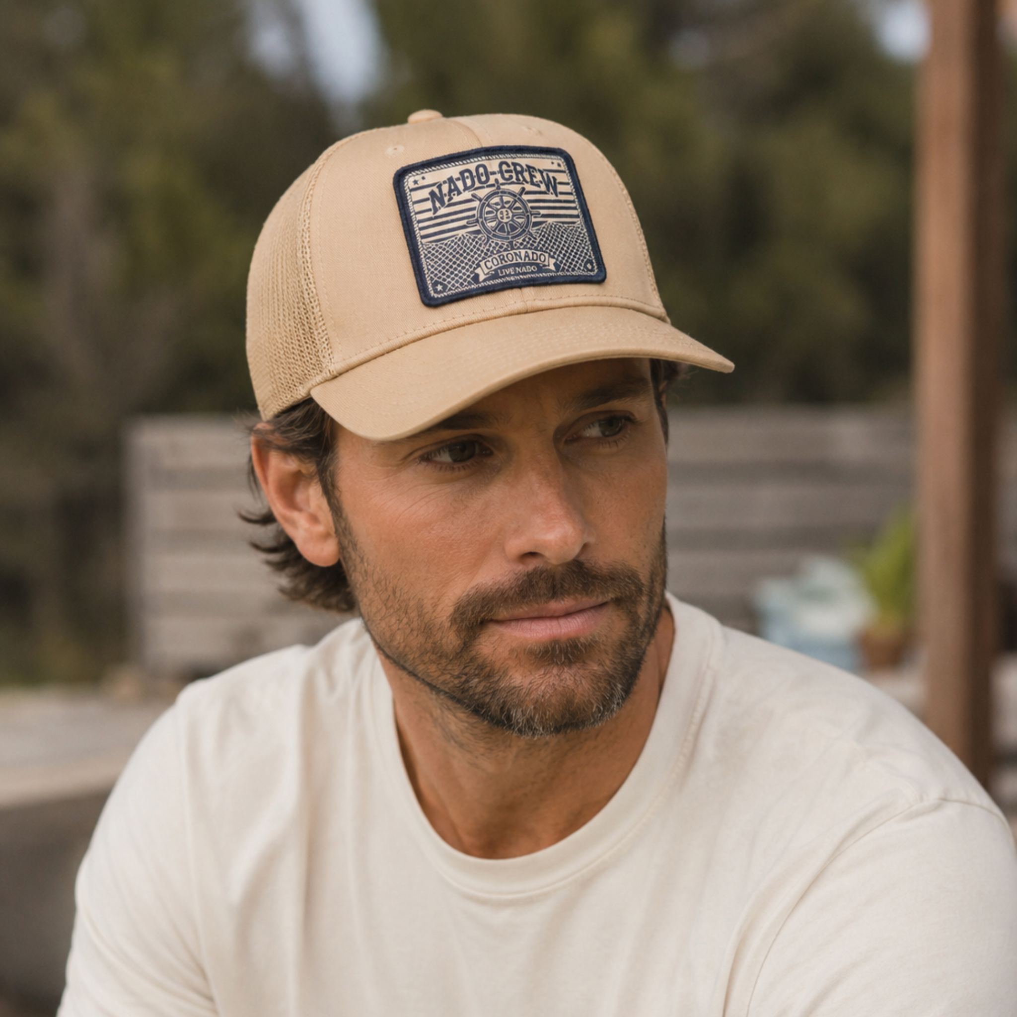 Man wearing a beige cap with a logo, sitting outdoors.