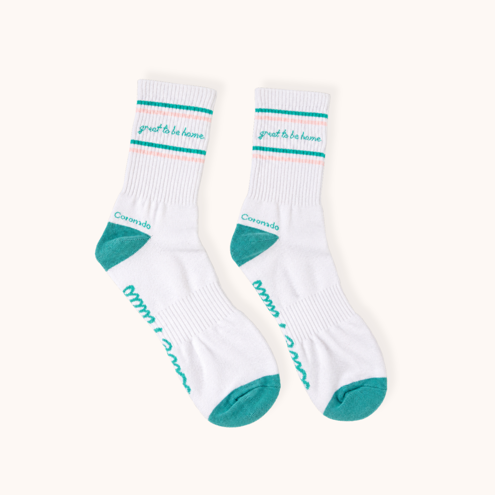 Flat lay view of a pair of white ribbed athletic crew socks on a light background. Each sock features teal and light pink double stripes near the top, with "great to be home." written in teal cursive between the stripes. The socks have teal heel caps, teal toe caps, and "Coronado" written above the heel.