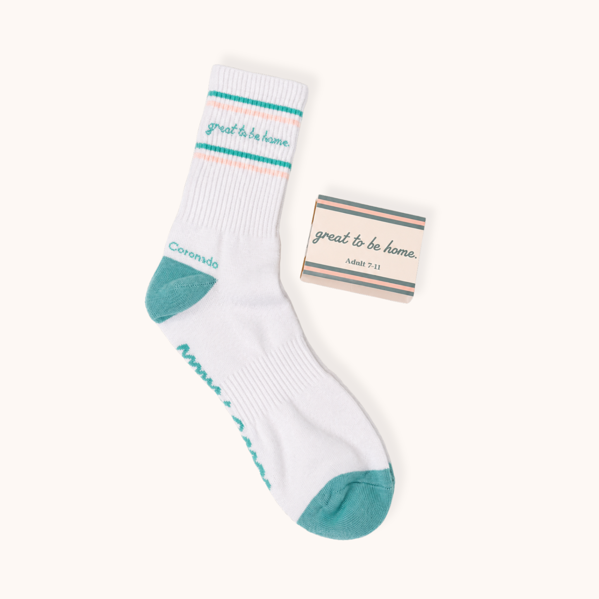Flat lay shot of a single white ribbed crew sock next to its rectangular kraft packaging tag. The sock features teal and light pink double stripes, teal cursive reading "great to be home.", teal heel and toe caps, and "Coronado" near the heel. The matching packaging sleeve displays the same cursive phrase, stripes, and size "Adult 7-11."