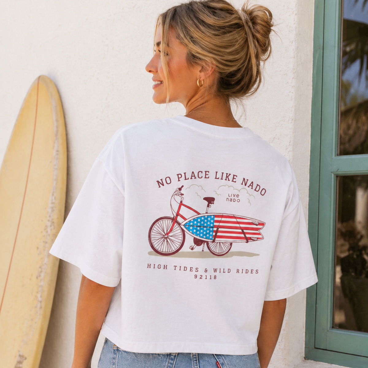 A woman wearing a white graphic t-shirt featuring a red bicycle carrying an American flag surfboard with the text "No Place Like Nado" and "High Tides & Wild Rides."