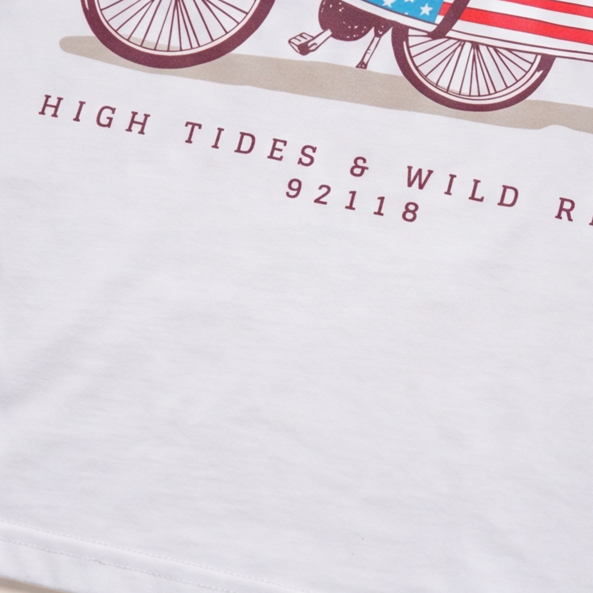 Extreme close-up of a white t-shirt showing graphic print detail reading "HIGH TIDES & WILD RIDES" and "92118" below a bicycle illustration.