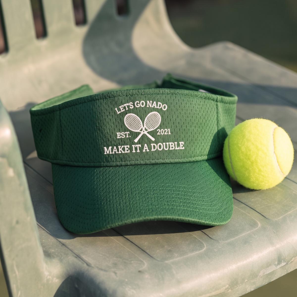 High-angle shot of a dark green athletic mesh visor resting on a light grey plastic court chair beside a bright yellow tennis ball. The front of the visor features white embroidered crossed tennis rackets, flanked by "EST. 2021," with "LET'S GO NADO" above and "MAKE IT A DOUBLE" below.