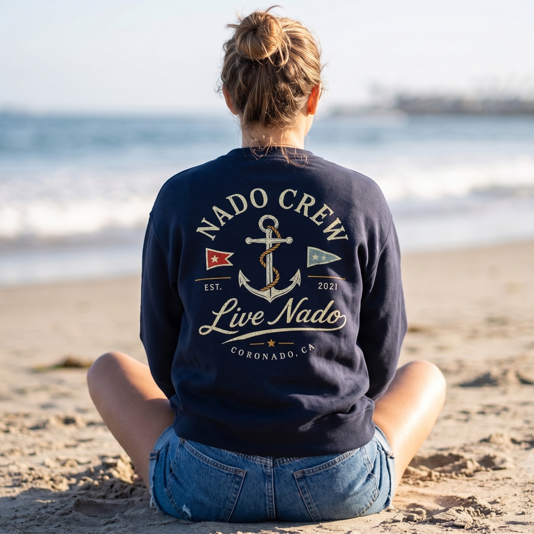 Back view of a woman with her hair in a bun sitting cross-legged in the sand facing the ocean. She is wearing denim shorts and a dark navy blue crewneck sweatshirt. The back of the sweatshirt features a vintage nautical design with an anchor, red and blue nautical flags, "NADO CREW" arched across the top, "Live Nado" in cursive, and "CORONADO, CA" written at the bottom.