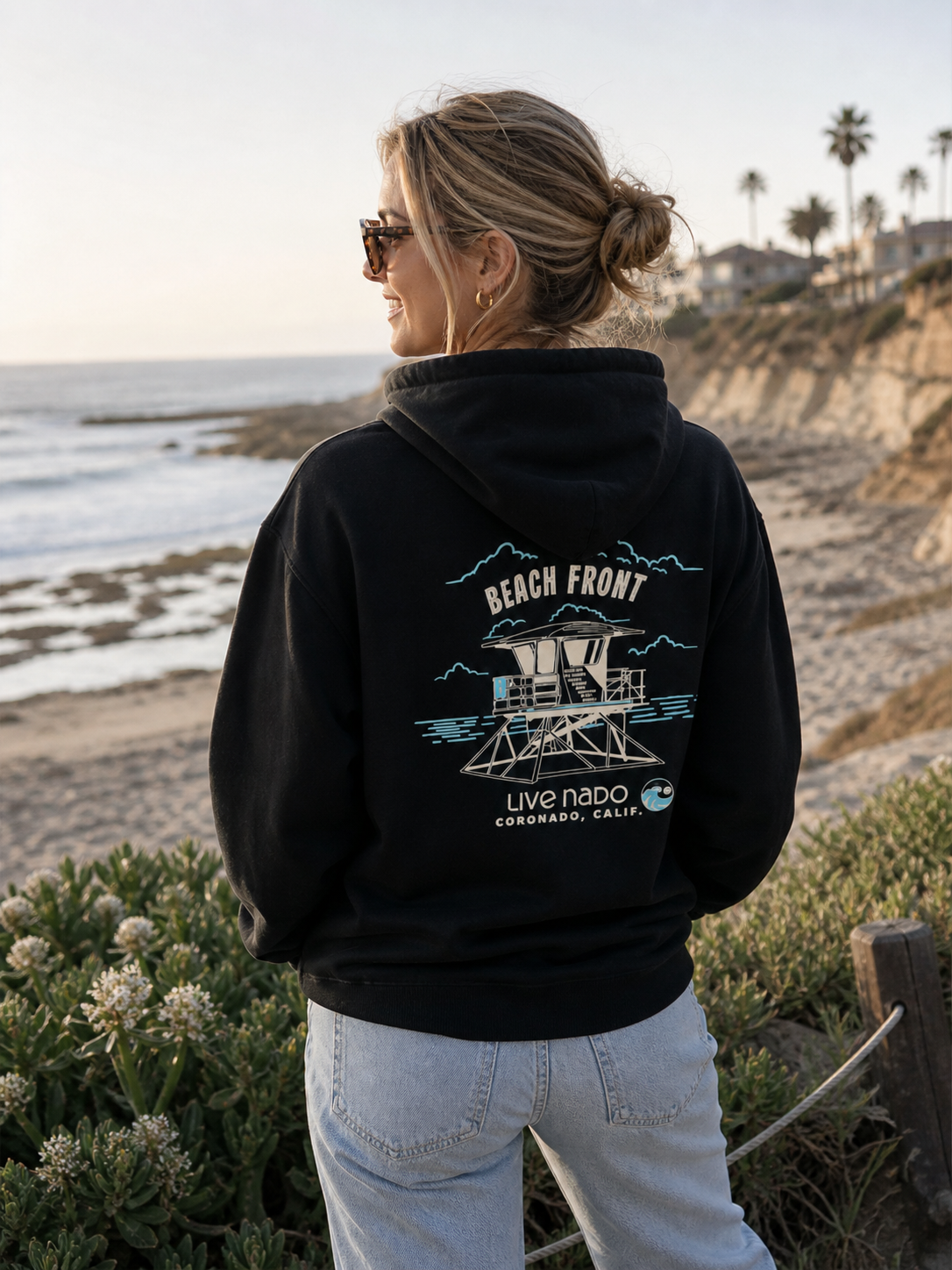 Woman wearing a black hoodie with a beach front design, standing on a beach.