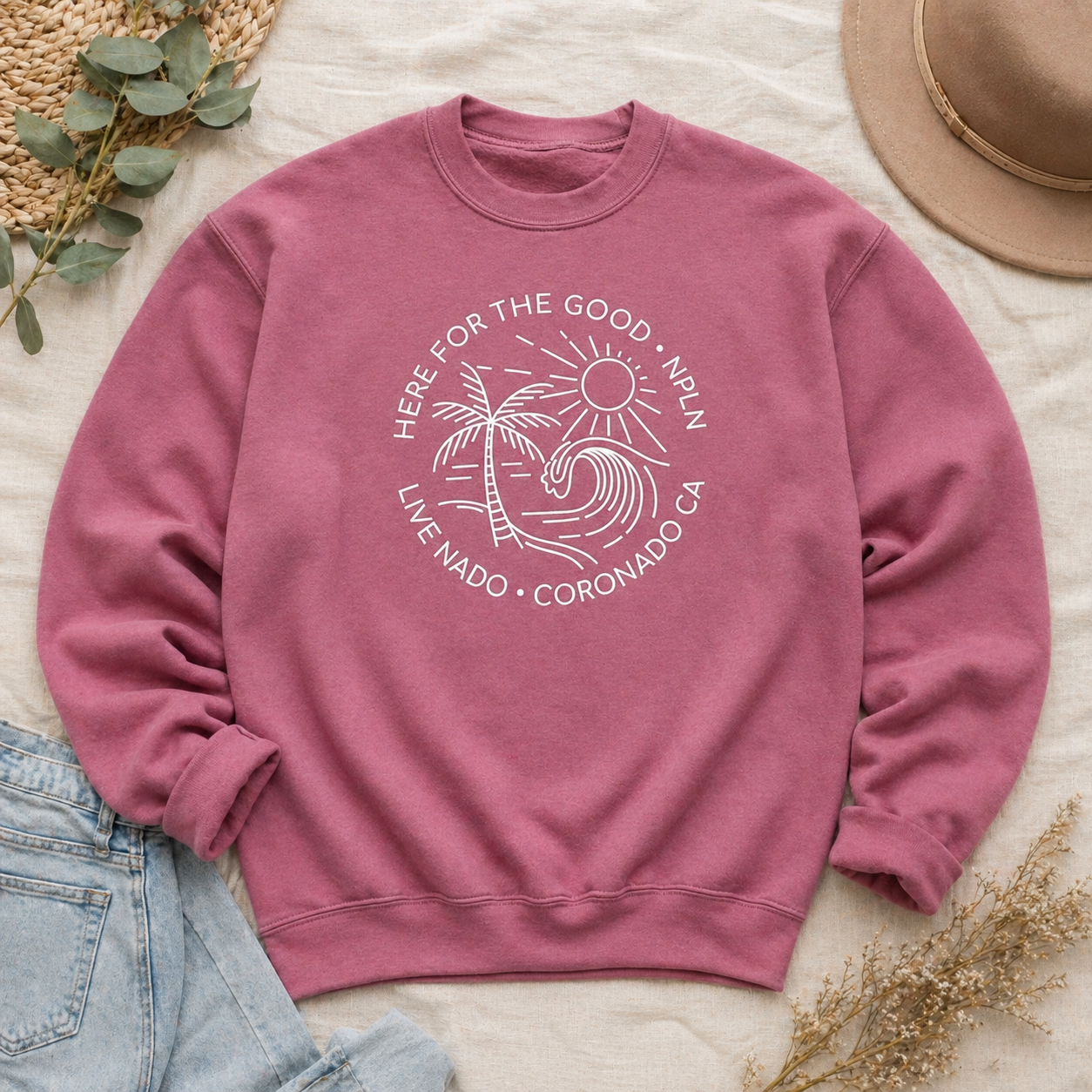 Pink sweatshirt with a palm tree design and text on a light surface with a hat, jeans, and leaves.