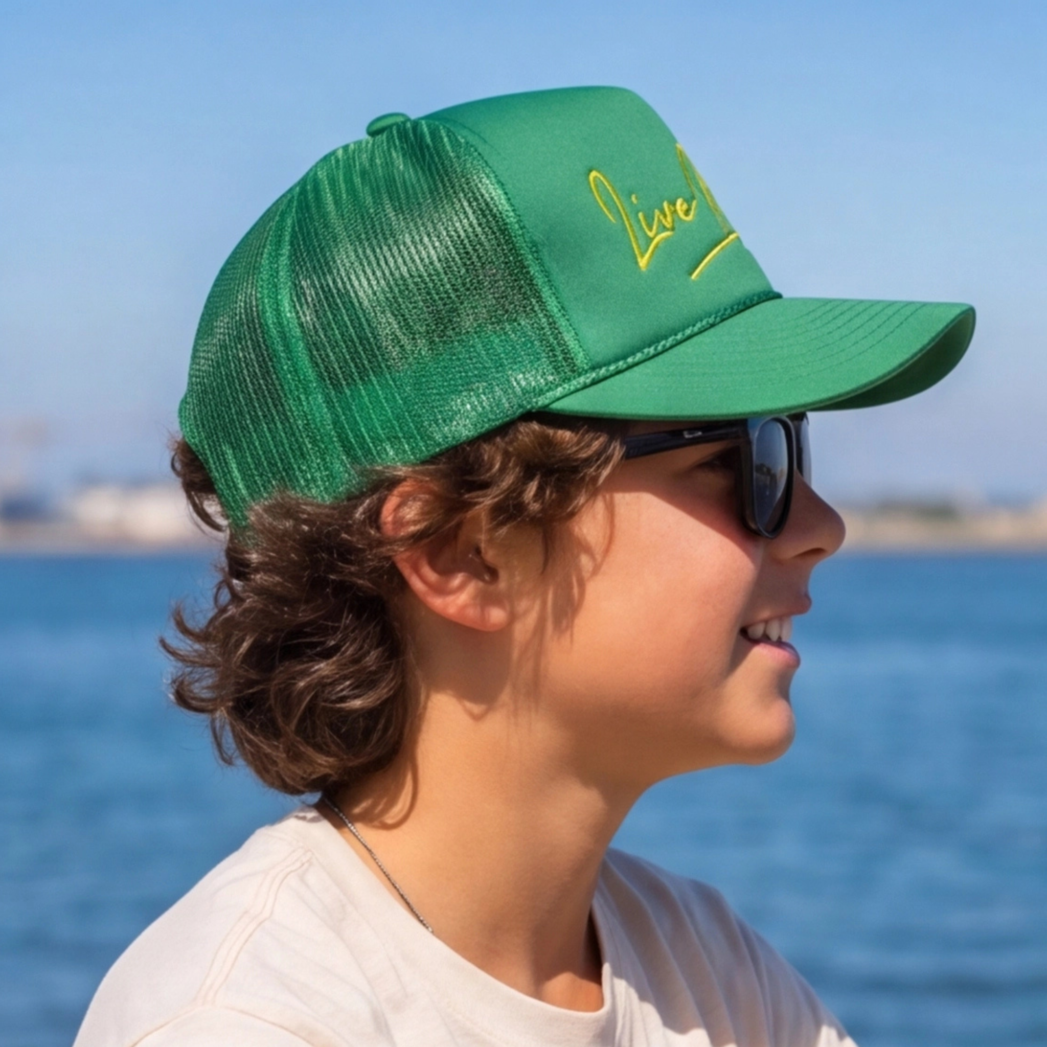 Child wearing a green cap with 'Live' text, sunglasses, and a white shirt by the water.