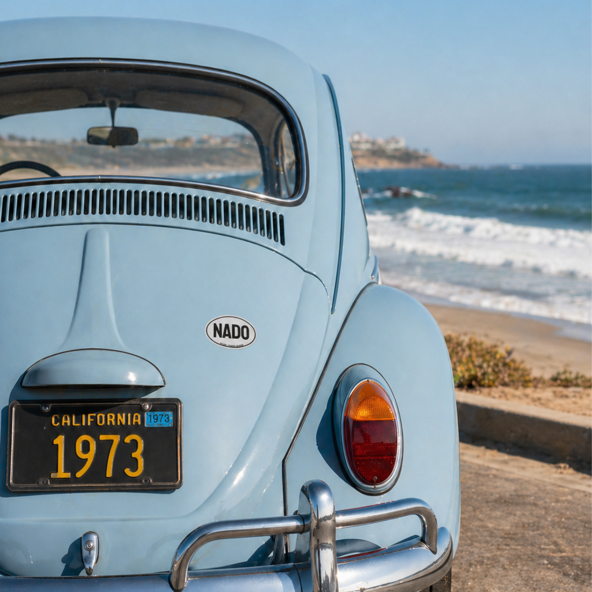 Vintage light blue car with 'NADO' logo and 'California 1973' license plate by the ocean.