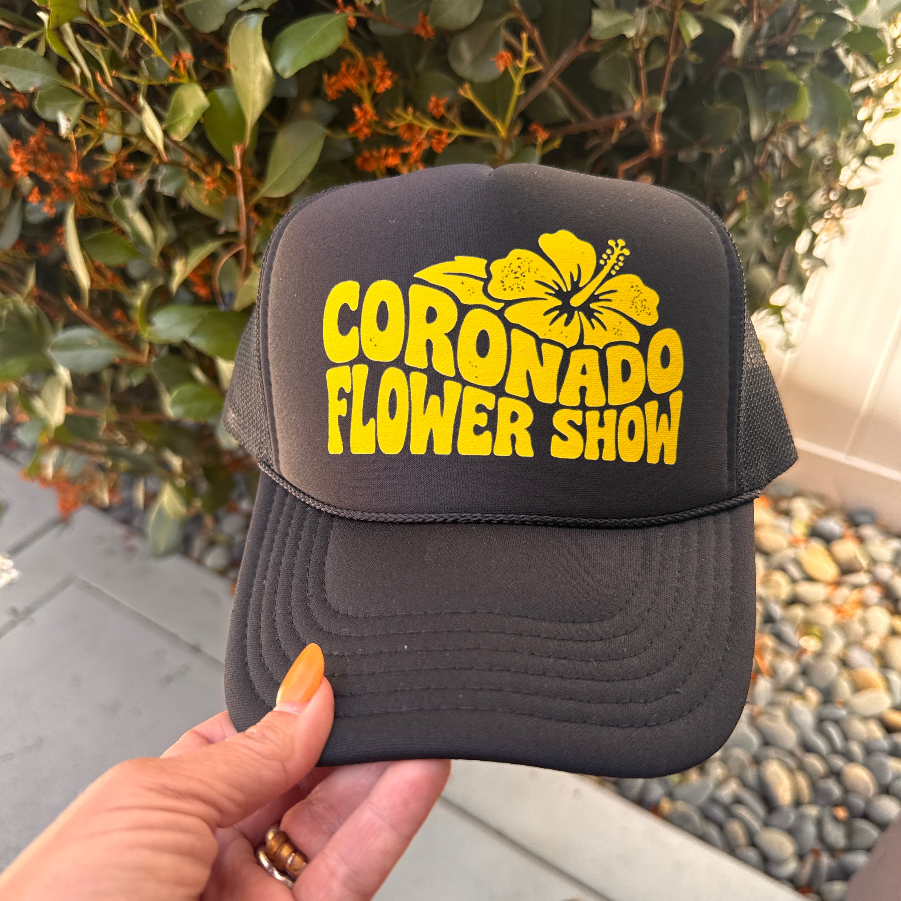Black cap with yellow 'Coronado Flower Show' text held by a hand against a natural background.
