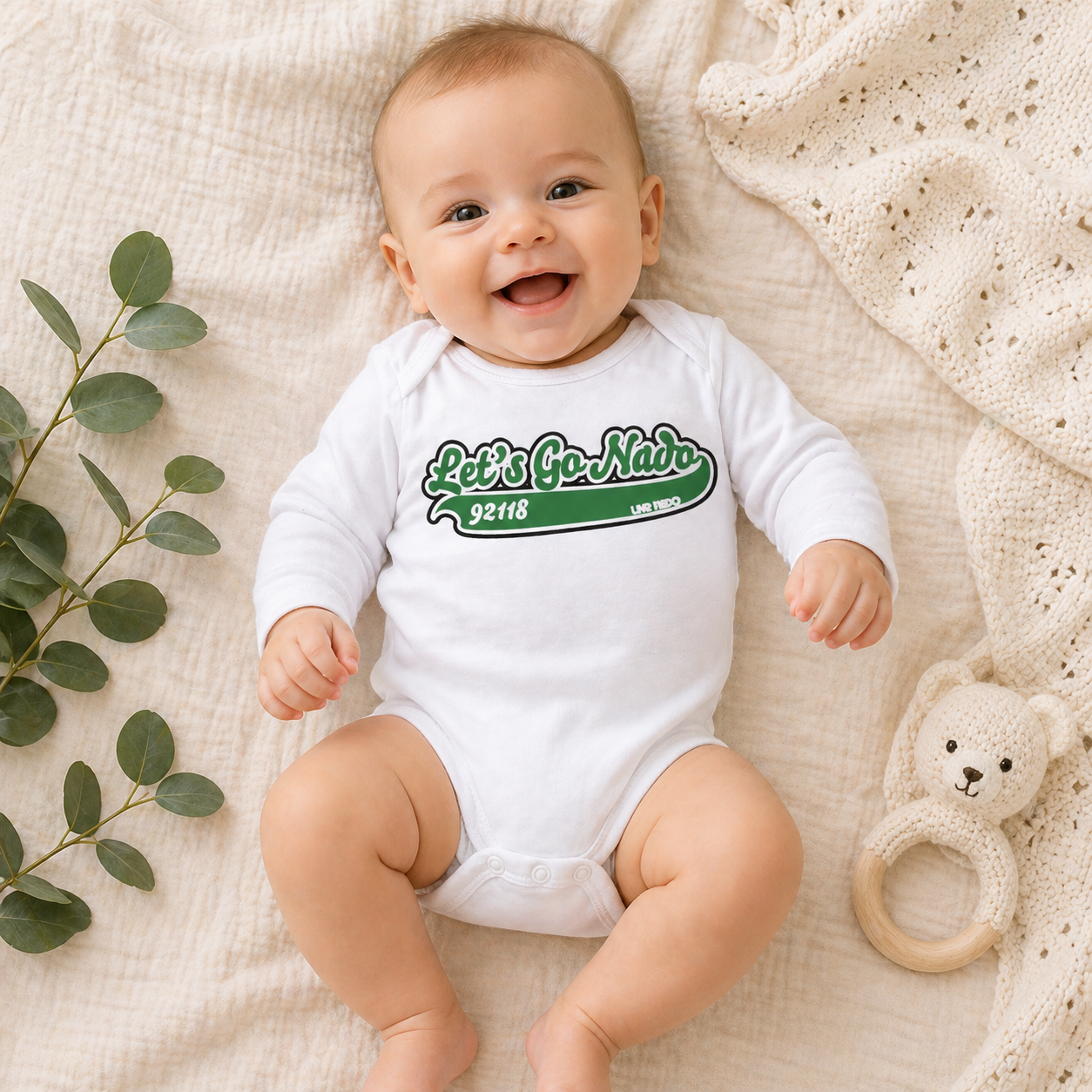 Baby wearing a white onesie with green text, lying on a textured surface with a plant and toy.
