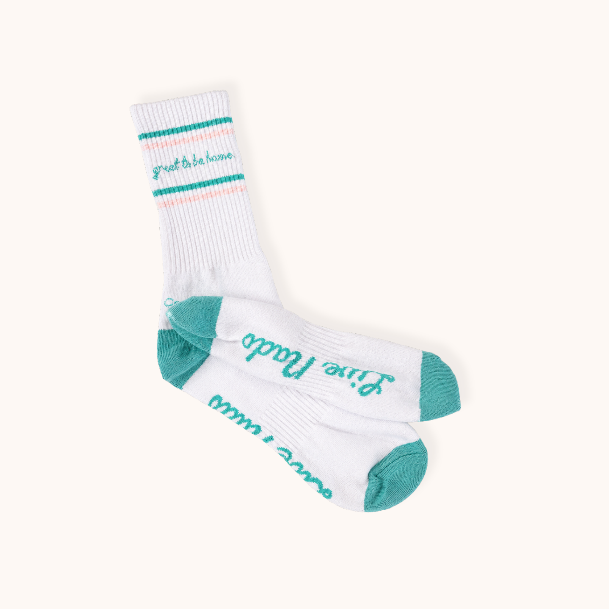 Flat lay view of a pair of white ribbed athletic crew socks crossed over one another on an off-white background. The upper cuff features teal and light pink double stripes with "great to be home." written in teal cursive. The soles display cursive teal lettering reading "Live Nado," complemented by teal heels and toes.