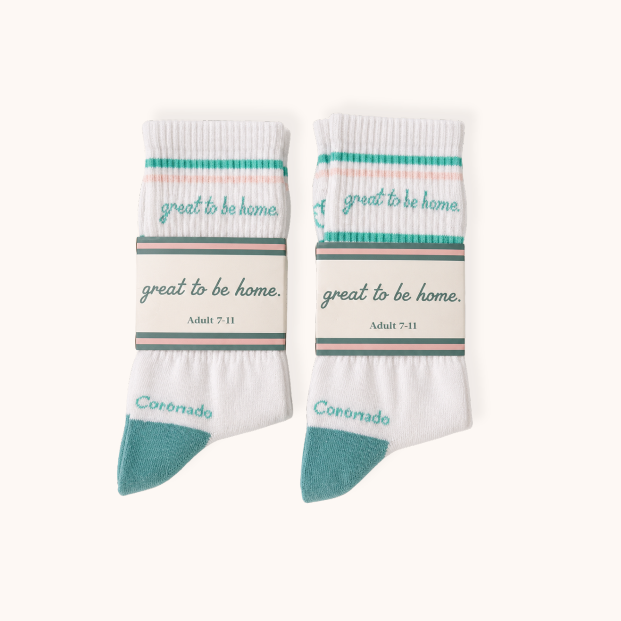 Flat lay view of two side-by-side folded pairs of white crew socks, each wrapped in a matching cardboard sleeve. The socks feature teal and pink stripes near the cuff, teal cursive reading "great to be home.", and teal heels with "Coronado" printed above. The packaging bands display the phrase "great to be home." and sizing "Adult 7-11."