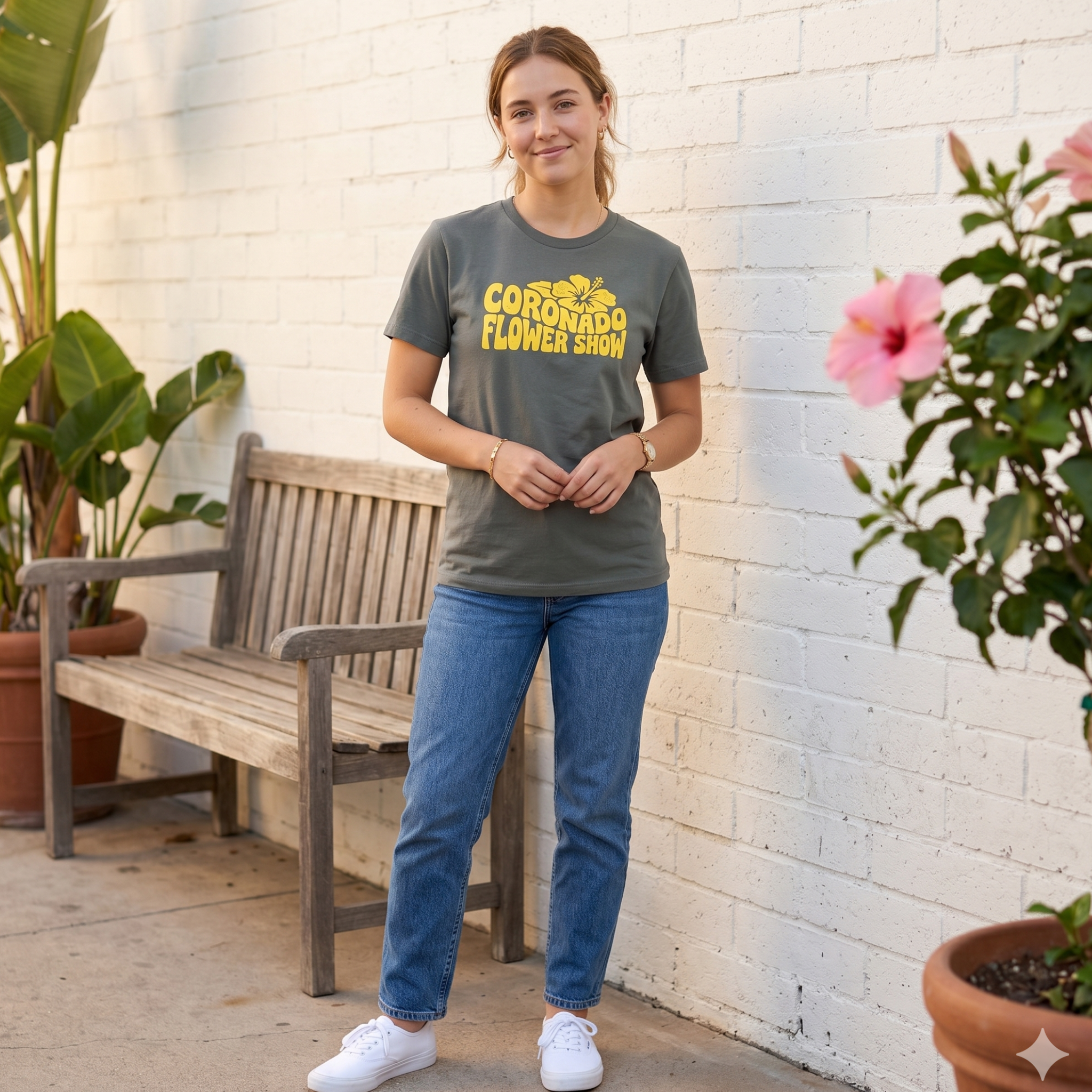 Person wearing a green t-shirt with 'Coronado Flower Show' text, standing outdoors near a wooden bench and plants.