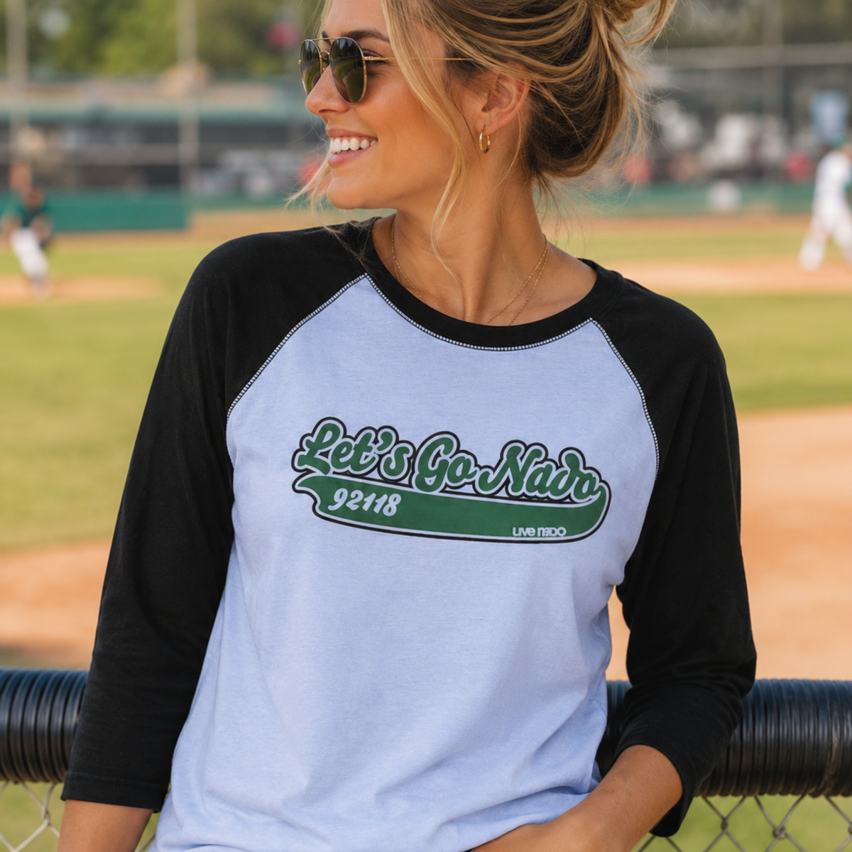 A smiling blonde woman with hair in a bun wearing sunglasses and gold hoop earrings. She is wearing a raglan-style shirt with black three-quarter sleeves and a light grey torso. The shirt features a green script graphic reading "Let's Go Nado" above a green banner with the zip code "92118." She leans against a outfield chain-link fence with a baseball field blurred in the background.