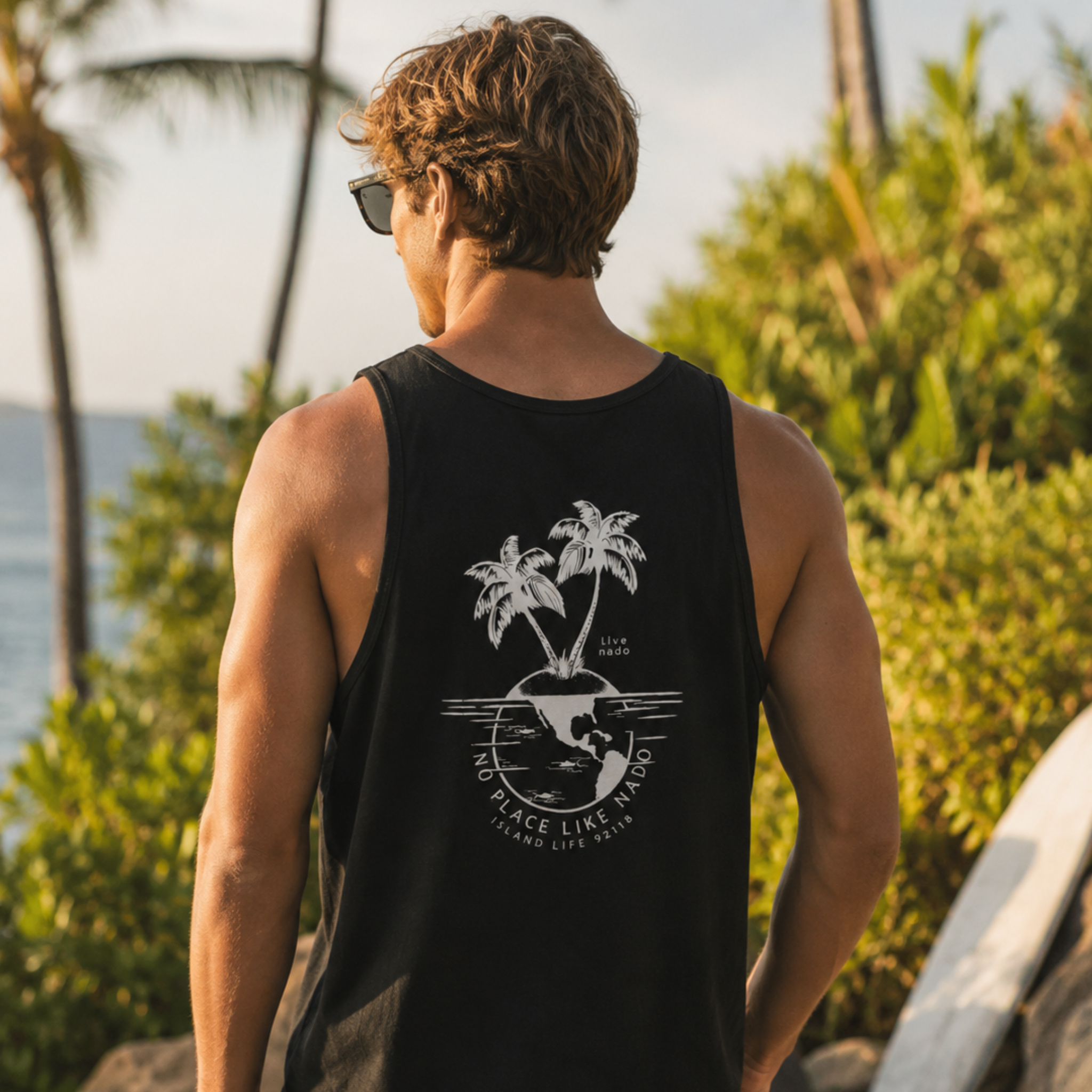 Back view of a wavy-haired man wearing sunglasses and a black tank top. The back of the tank features a white graphic illustration of two palm trees growing on top of a globe surrounded by the text "NO PLACE LIKE NADO" and "ISLAND LIFE 92118." He stands outdoors facing away from the camera, with tropical palm trees and the ocean blurred in the background.