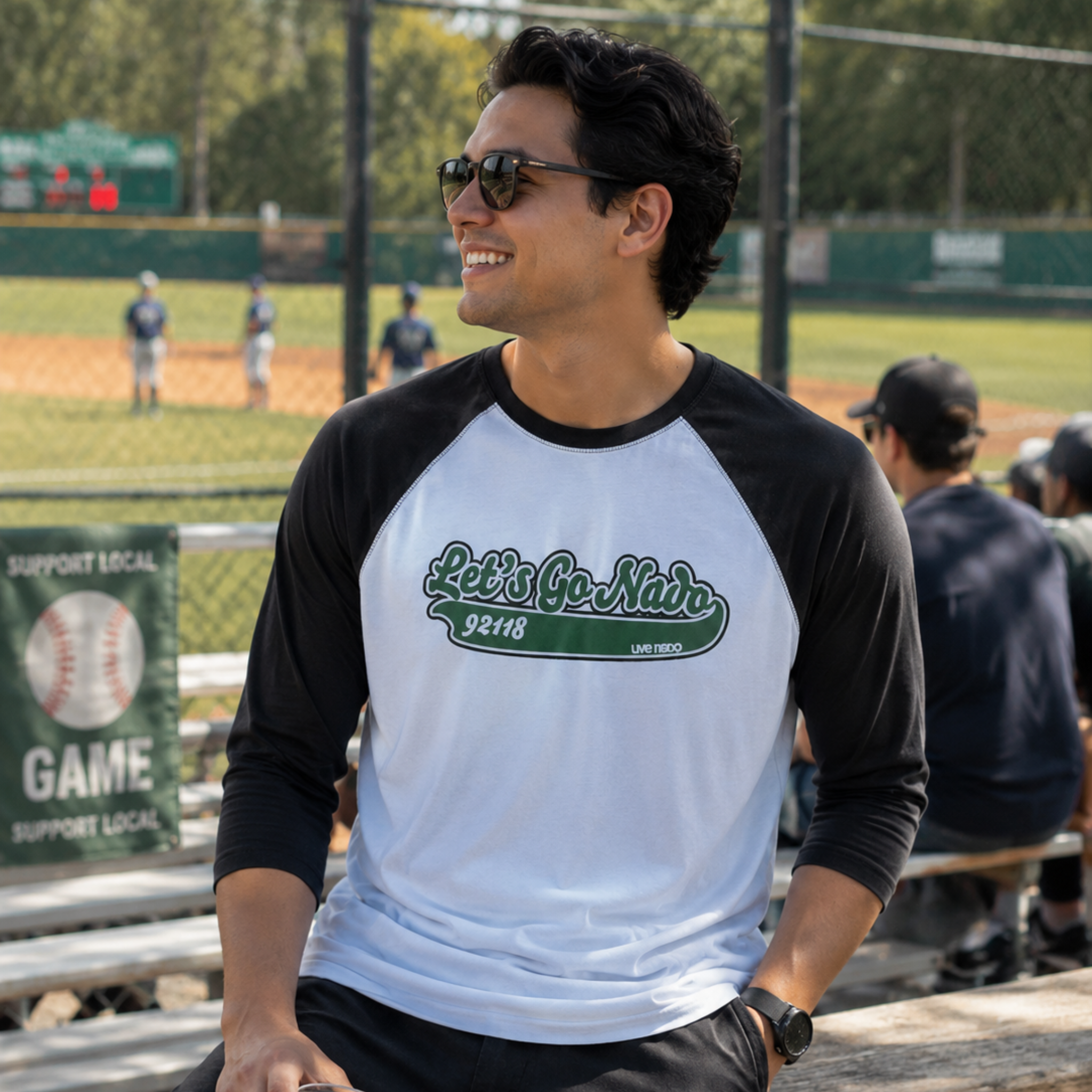 A dark-haired man with dark sunglasses smiling while looking off-camera. He is sitting on wooden bleachers at a baseball stadium wearing a raglan-style shirt with black three-quarter sleeves and a light grey torso. The chest features a green cursive graphic reading "Let's Go Nado 92118." A baseball field with players and a fence is softly blurred in the background.