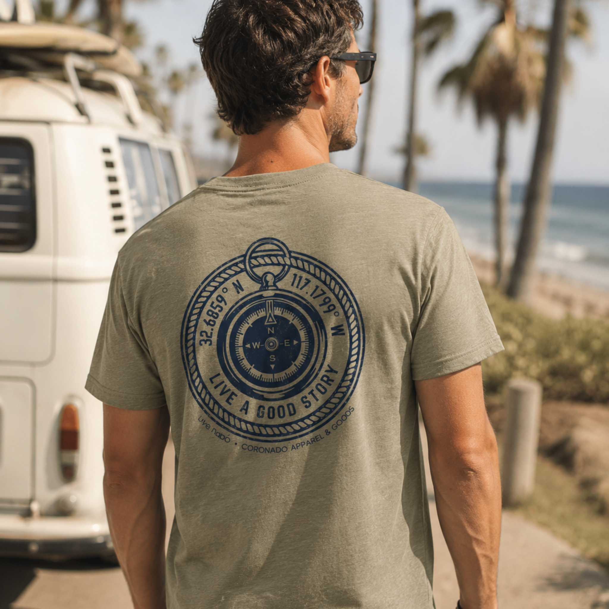 Rear profile of a man wearing sunglasses and a heather sage green t-shirt featuring a large dark blue circular compass graphic on the back. The graphic includes coordinates and the phrase "LIVE A GOOD STORY." He stands outdoors next to a vintage white camper van overlooking a tropical beach with palm trees.