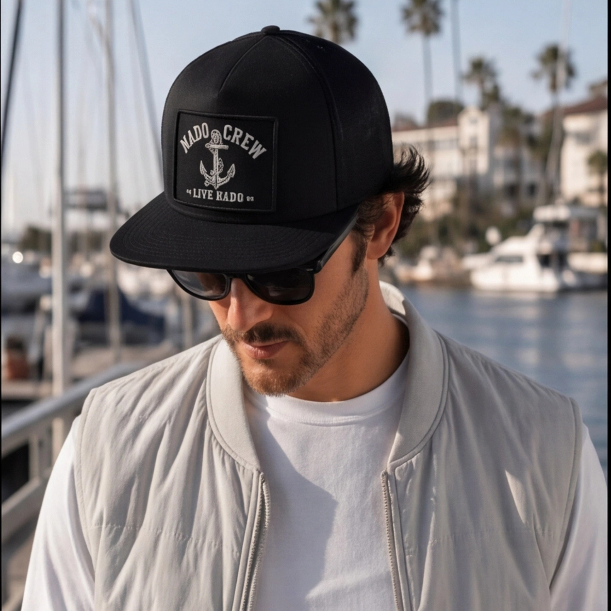 Man wearing a black cap with branding and sunglasses, standing by a waterfront with boats and palm trees.