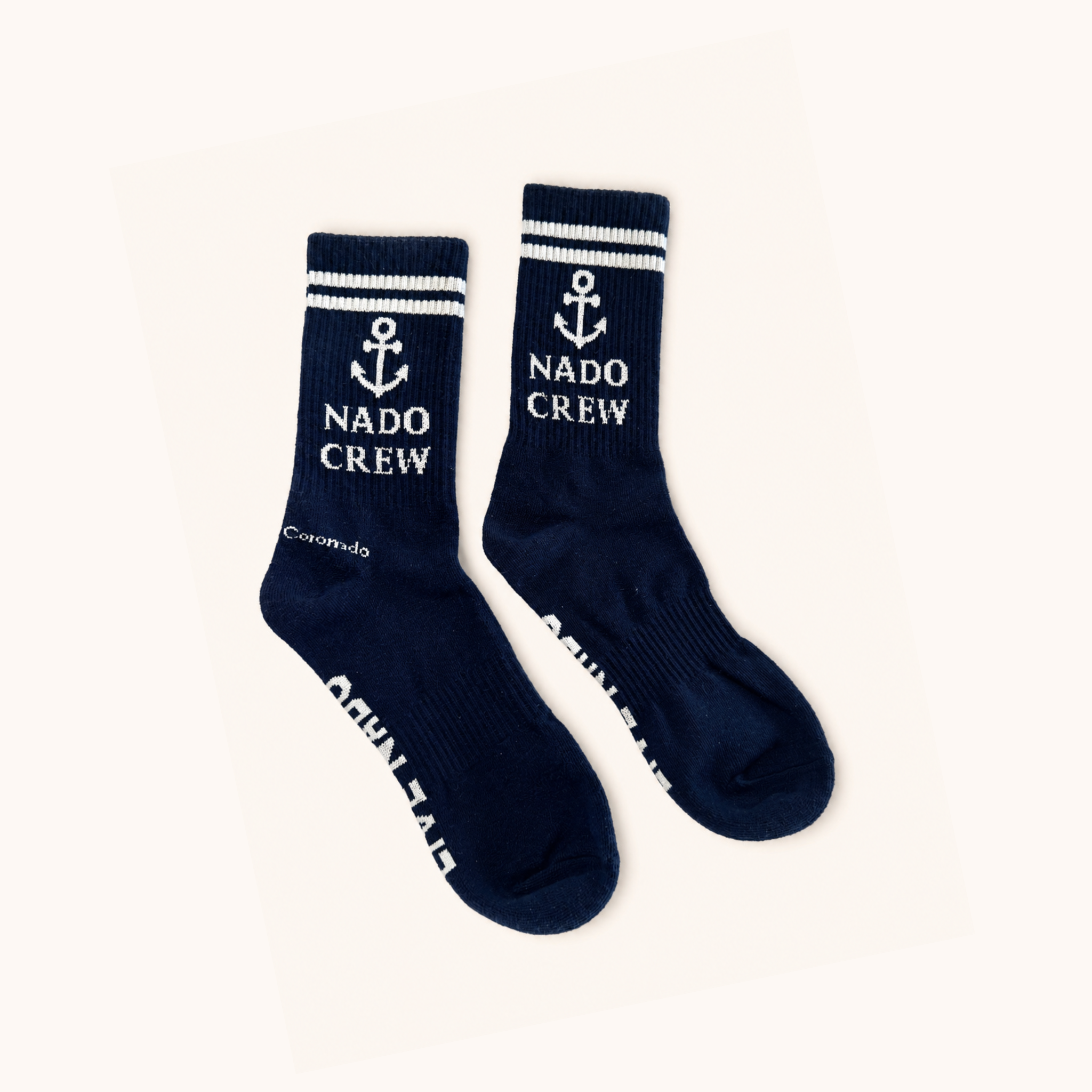 Flat lay view of a pair of dark navy blue ribbed crew socks laid against a off-white background. The top cuffs feature double white athletic stripes above a white anchor emblem and the text "NADO CREW." Small white text reading "Coronado" is near the heel, and text is knitted along the sole.