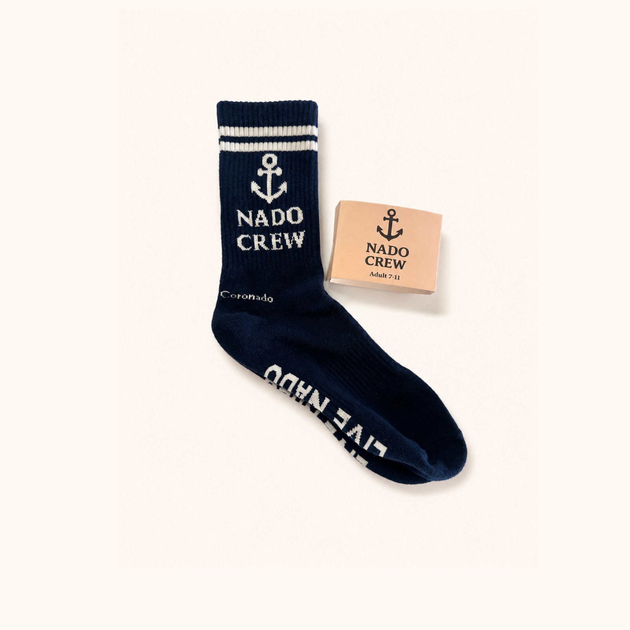 Flat lay shot of a single navy blue ribbed crew sock against a off-white background. The sock features double white stripes at the top, a white anchor icon, and "NADO CREW" text on the leg. "Coronado" is written near the heel, and "LIVE NADO" is knitted on the sole. A kraft paper packaging sleeve reading "NADO CREW Adult 7-11" sits beside it.