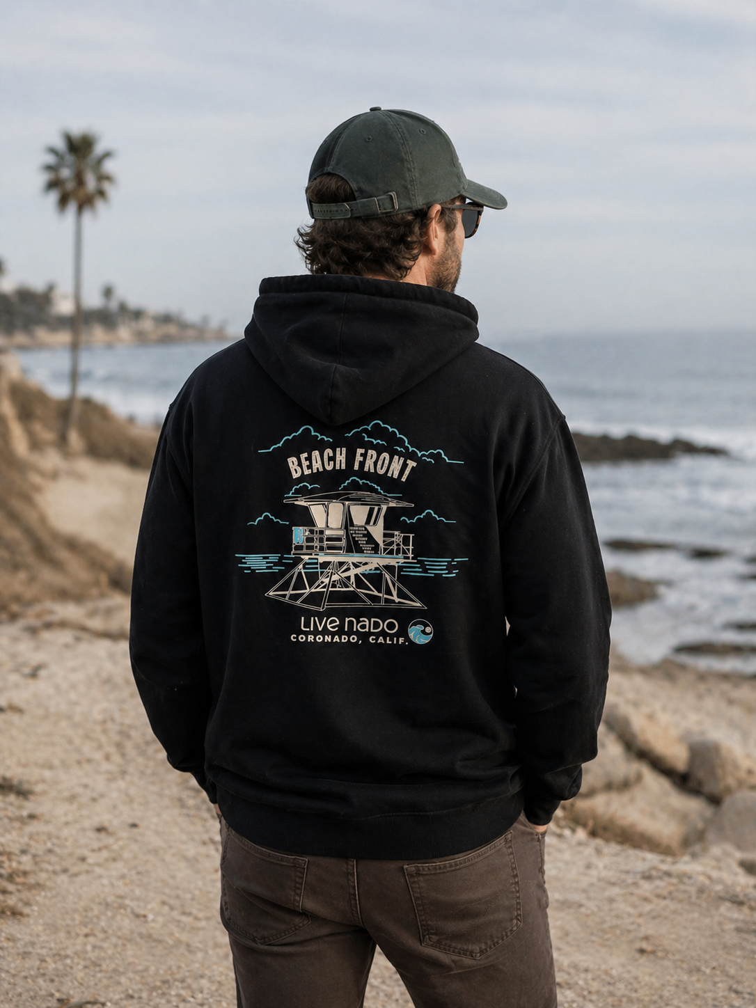 Person wearing a black hoodie with a beach front design, standing on a coastal path.