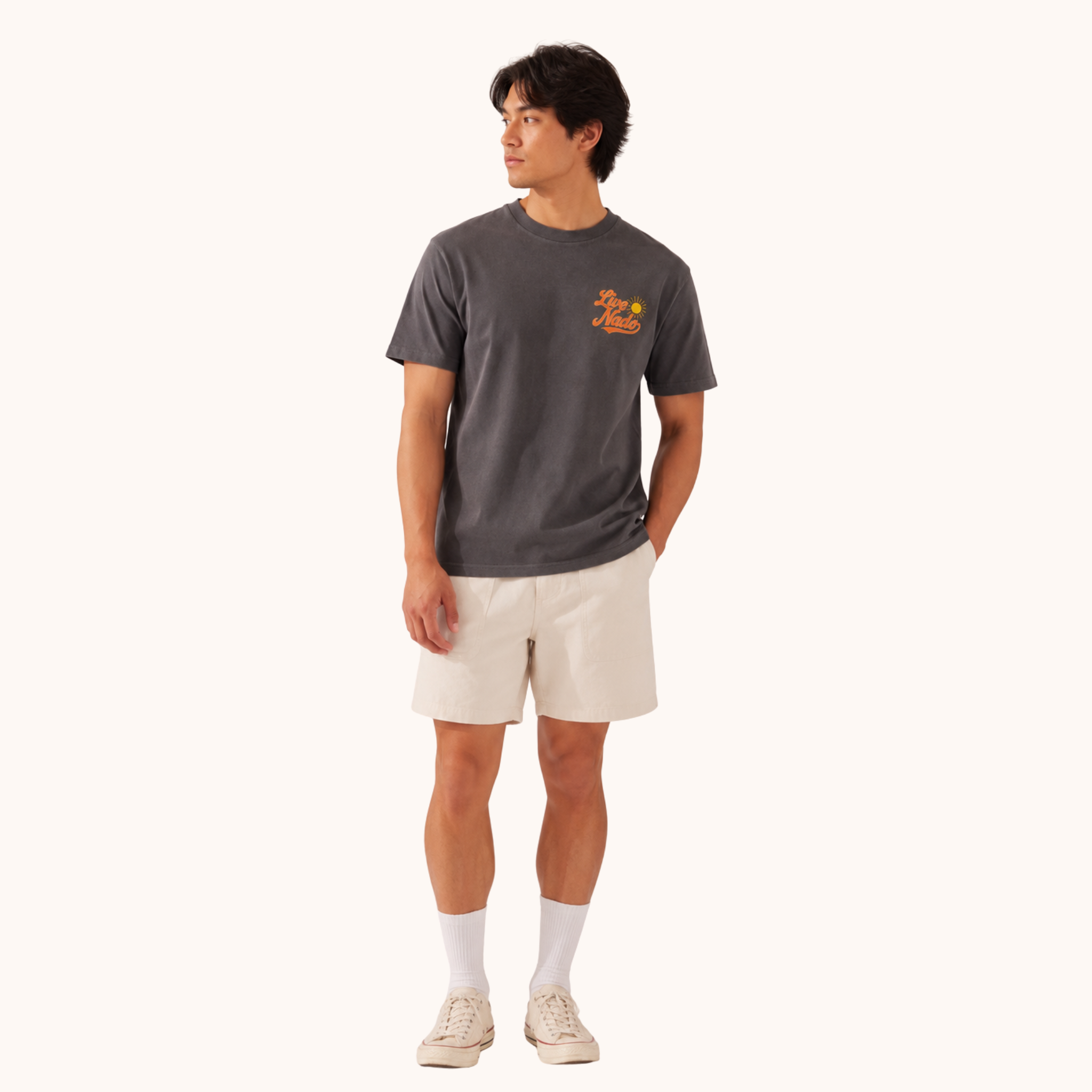 Man wearing a charcoal gray t-shirt with an orange "Live Nado" logo on the left chest, paired with off-white shorts and white sneakers.